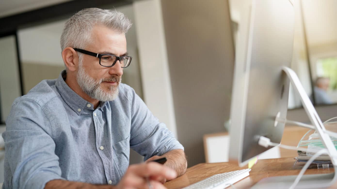 Man with grey hair and glasses works on graphic design project on computer screen.